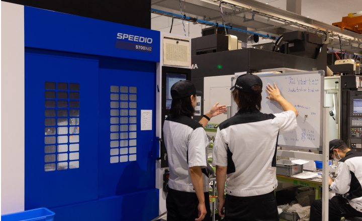 工作機械の前でホワイトボードを指し示しながら打ち合わせをする社員