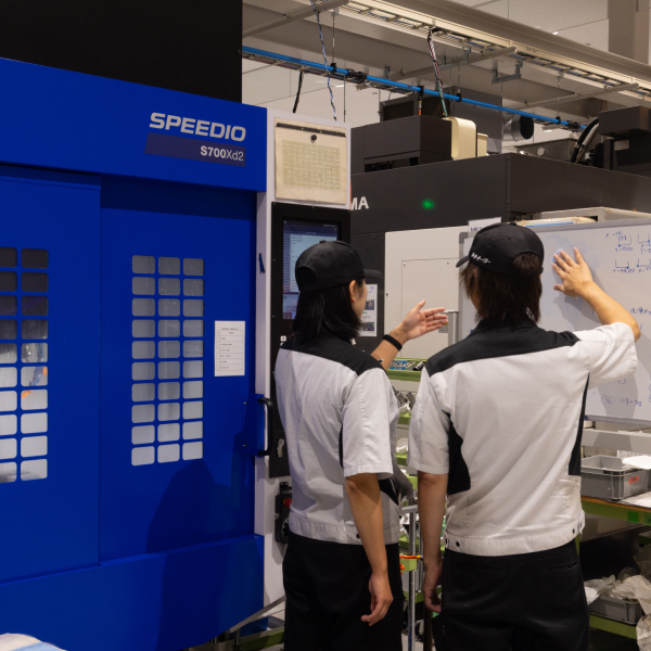工作機械の前でホワイトボードを指し示しながら打ち合わせをする2人の社員