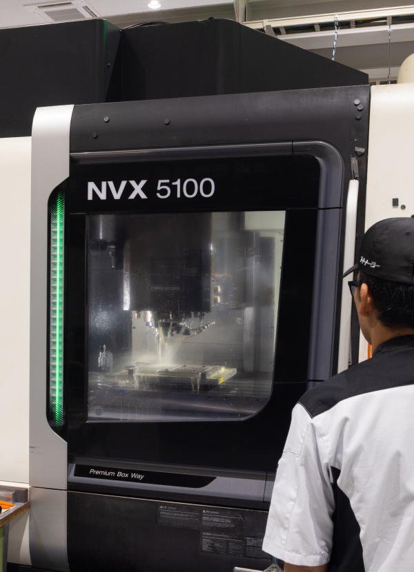 工作機械の内部で切削液をかけながら金属部品が加工されている様子を確認する社員